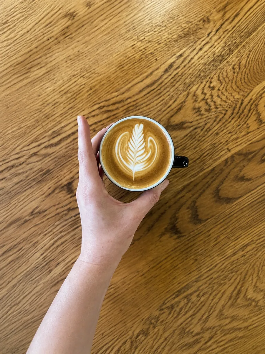 A cup of coffee with leaf-shaped latte art, held over a timber floor